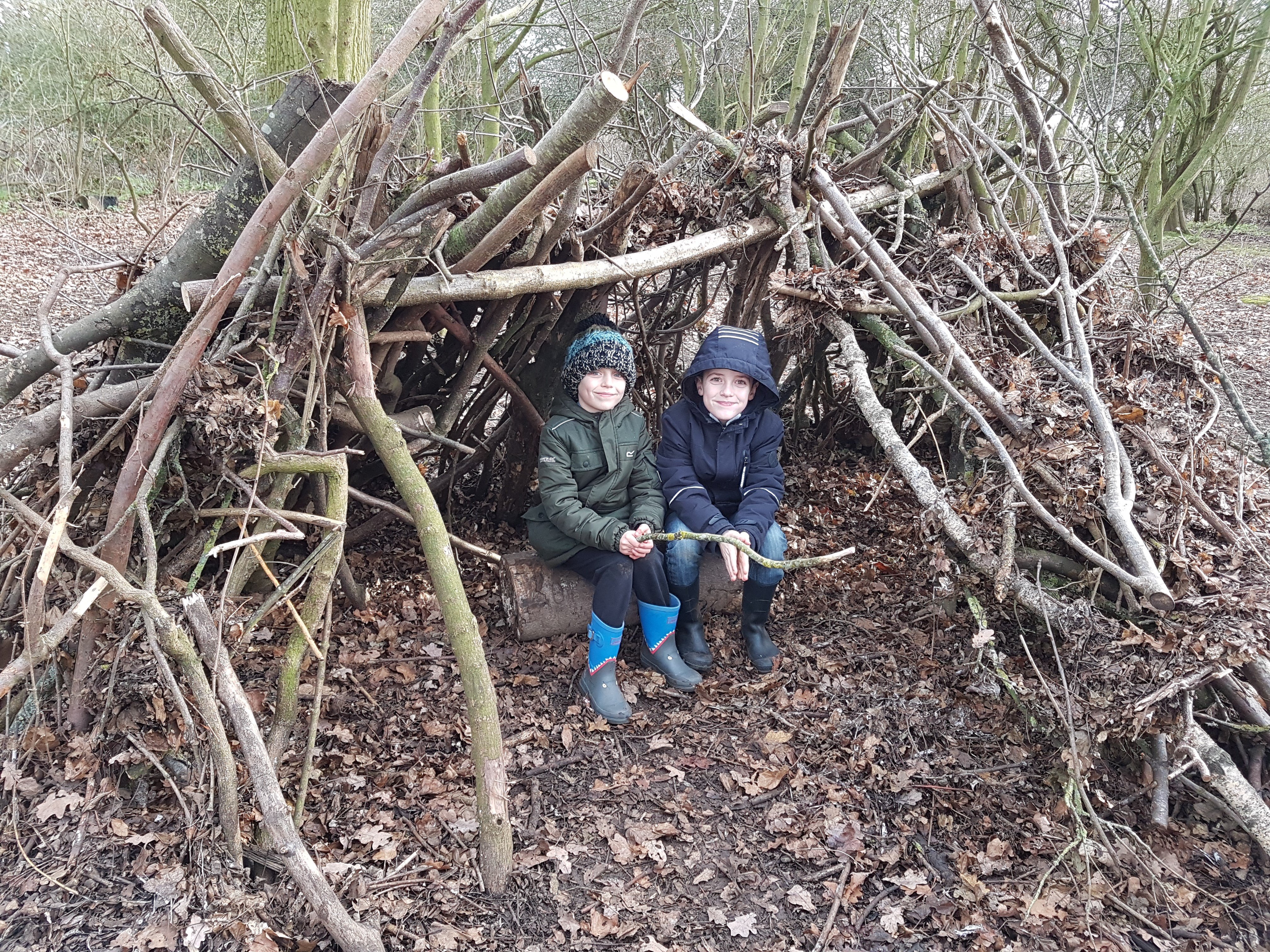 Two young boys sat in a shelter built from sticks in a wood